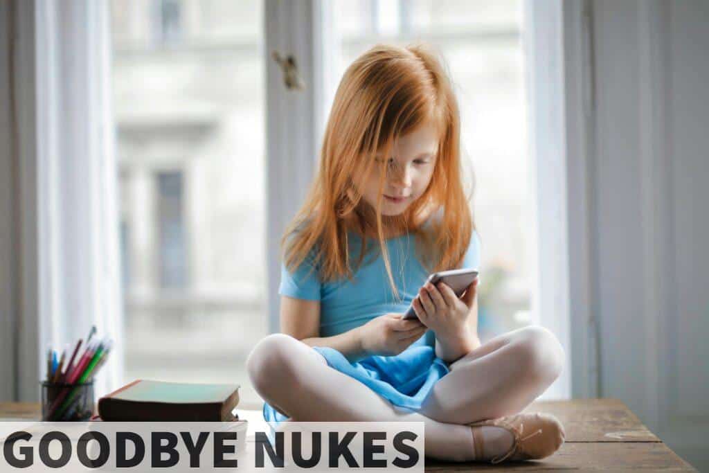 Red haired charming schoolgirl in blue dress browsing smartphone while sitting on rustic wooden table with legs crossed beside books against big window at home