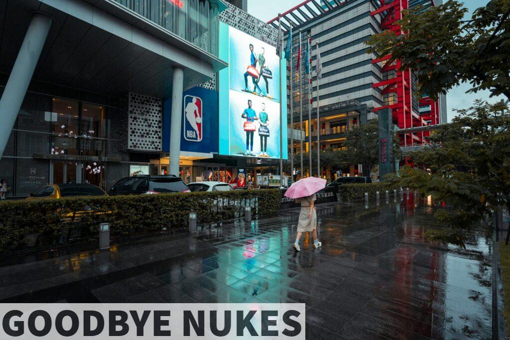 A woman walks with an umbrella past modern urban buildings and wet pavement reflecting city lights.